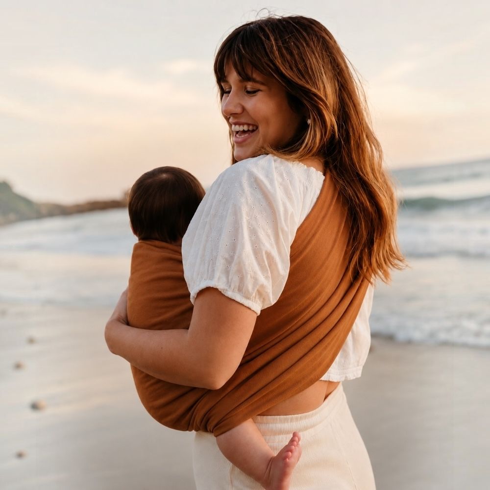 Anne, bebeğini kahverengi halka sling ile güvenli ve ergonomik şekilde taşırken sahilde yürümektedir. Bebek arabasını açmadan pratik bebek taşıma çözümü sunan halka sling, günlük yaşamda konforlu ve eller serbest kullanım sağlar.