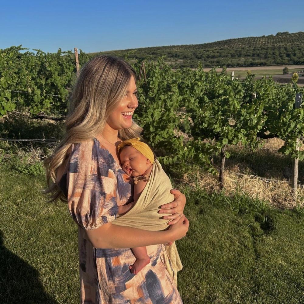 Doğal ortamda ergonomik halka sling ile bebeğini taşıyan anne. Ergonomik bebek taşımanın omurga gelişimi, kalça desteği, güvenli bağlanma ve bebeğin fiziksel gelişimine katkılarını anlatan yaşam fotoğrafı.