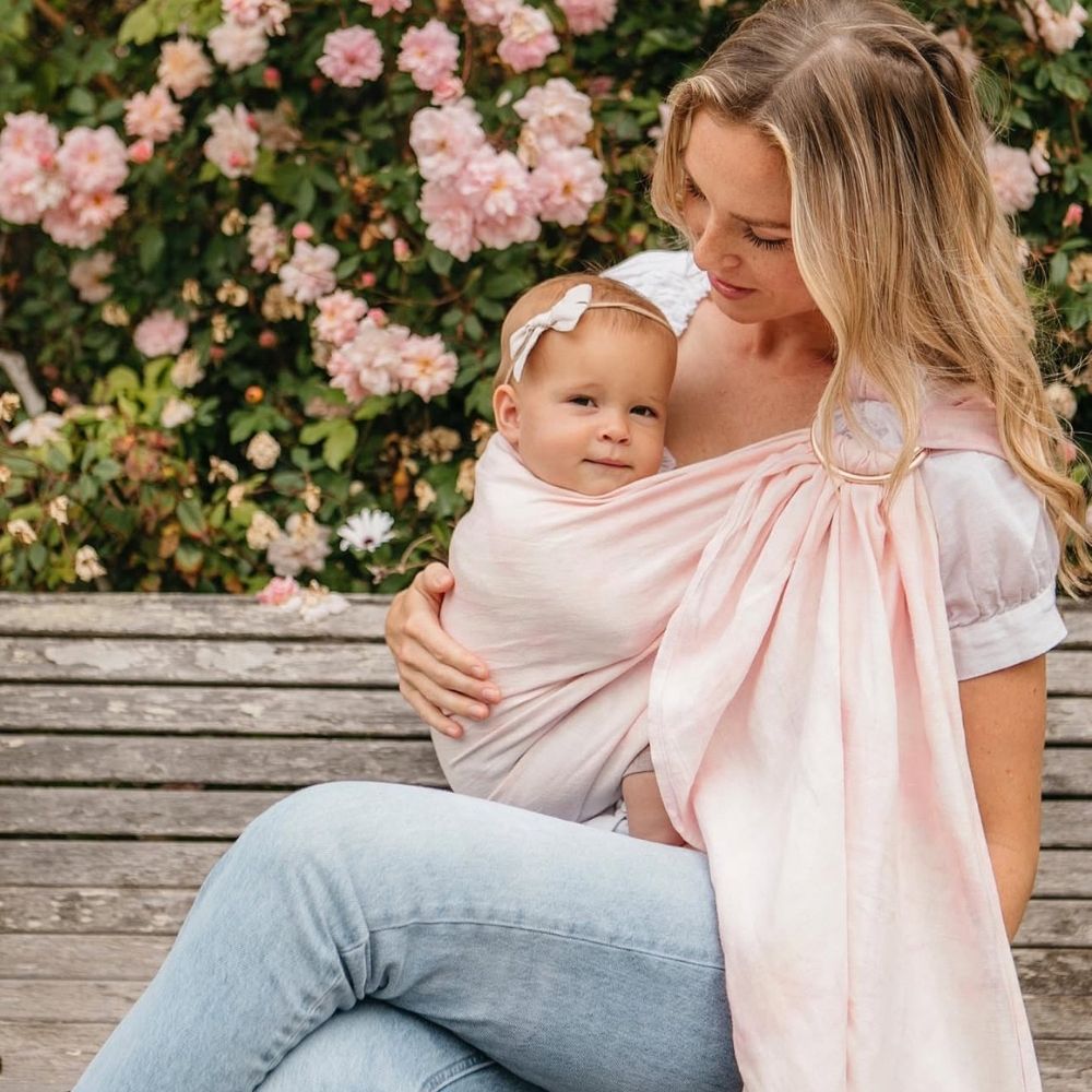 Pembe halka sling içinde bebeğini güvenli şekilde taşıyan anne. Halka sling alırken yapılan yaygın hataları önlemeye yardımcı olacak doğru kumaş, ergonomik kullanım ve güvenli taşıma örneğini gösteren yaşam fotoğrafı.