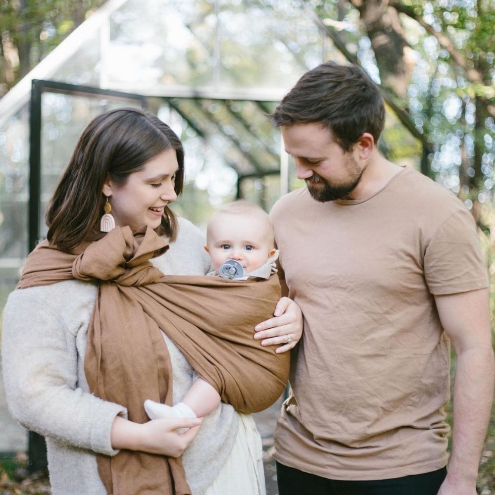 Anne, baba ve bebek kahverengi halka sling ile bir arada. Halka sling, yeni anne ve babalar için hem anlamlı hem de uzun süre kullanılabilen ergonomik bir bebek taşıma hediyesidir.