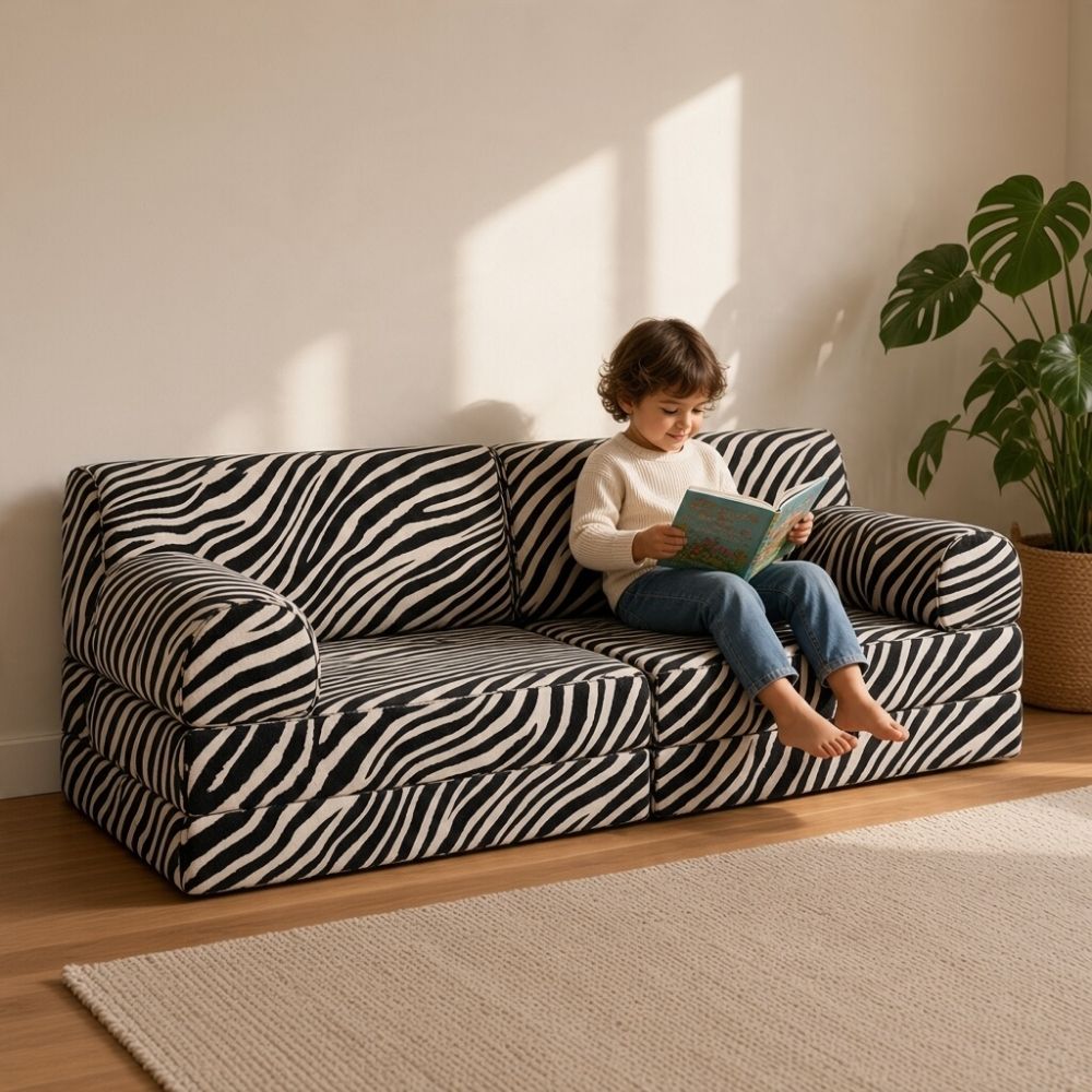 Küçük bir salonda siyah beyaz zebra desenli modüler çocuk koltuğunda kitap okuyan çocuk. Az yer kaplayan, çok amaçlı çocuk koltuğu kullanımına örnek.
