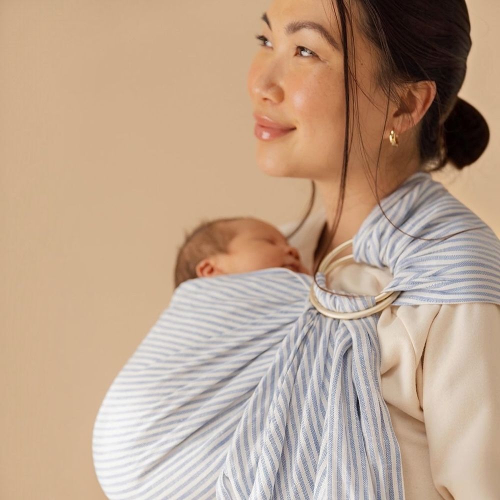 Anne, yenidoğan bebeğini mavi çizgili halka sling ile ergonomik ve güvenli şekilde taşıyor.